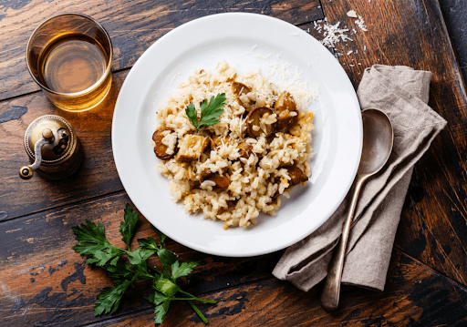 Fresh Porcini mushrooms used in a Risotto, on a white plate with a spoon to the right, garnish underneath, and a cup of water on the wood table.