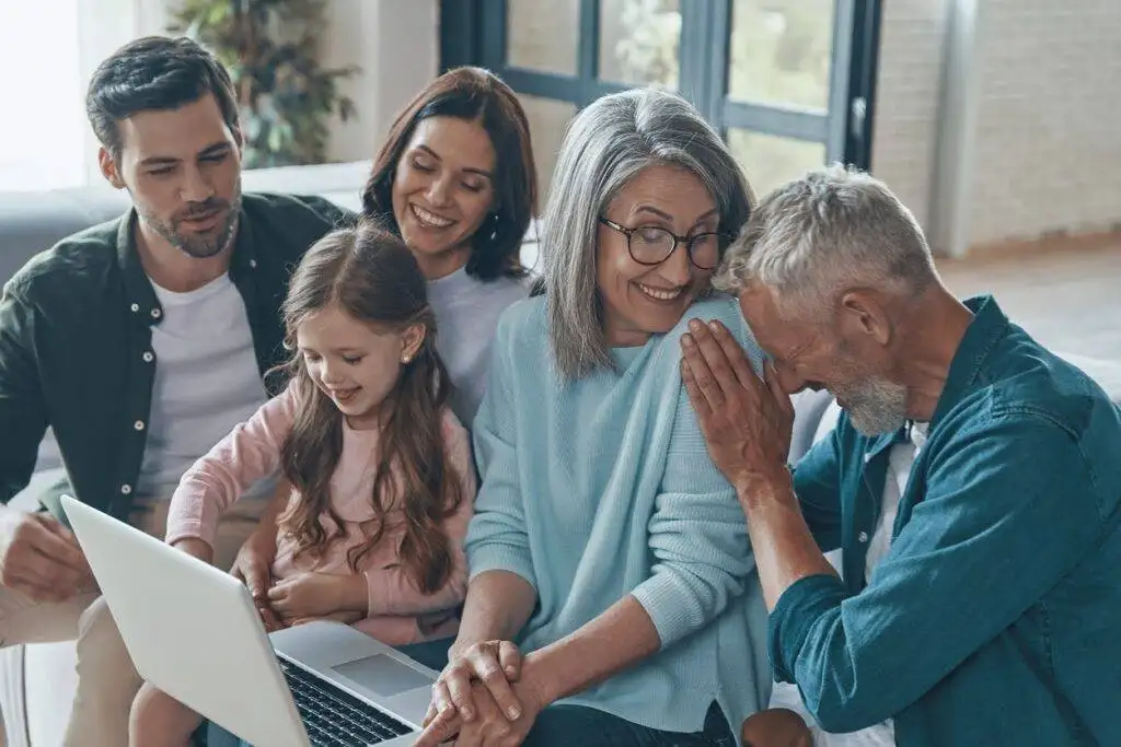 Husband and wife sitting on a couch with their young daughter on their lap and the grandparents also sitting on the couch to their left, everyone is laughing.